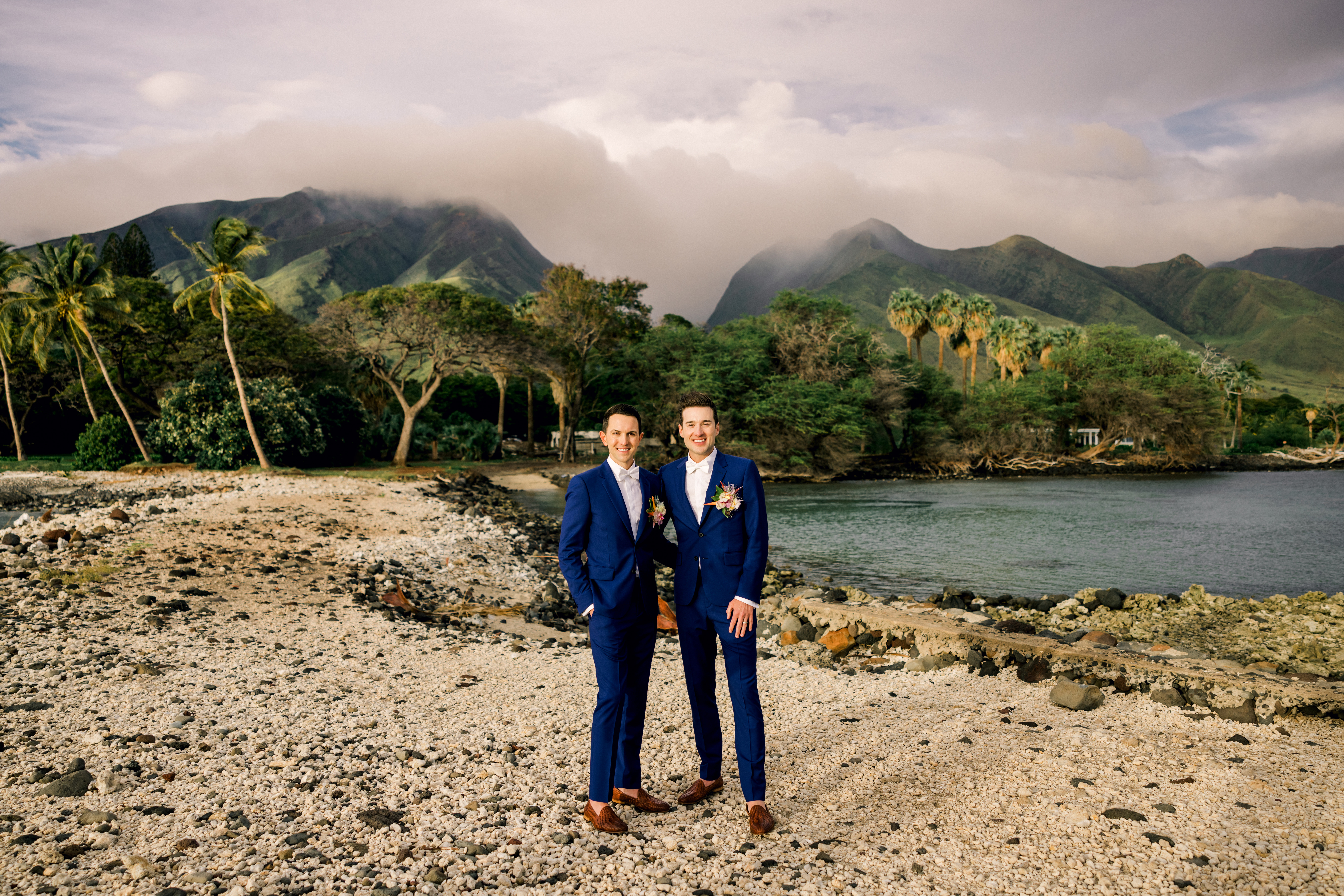 Newlywed portraits standing side by side in matching suit, bow tie, and shoes with the ocean and mountains in the background.