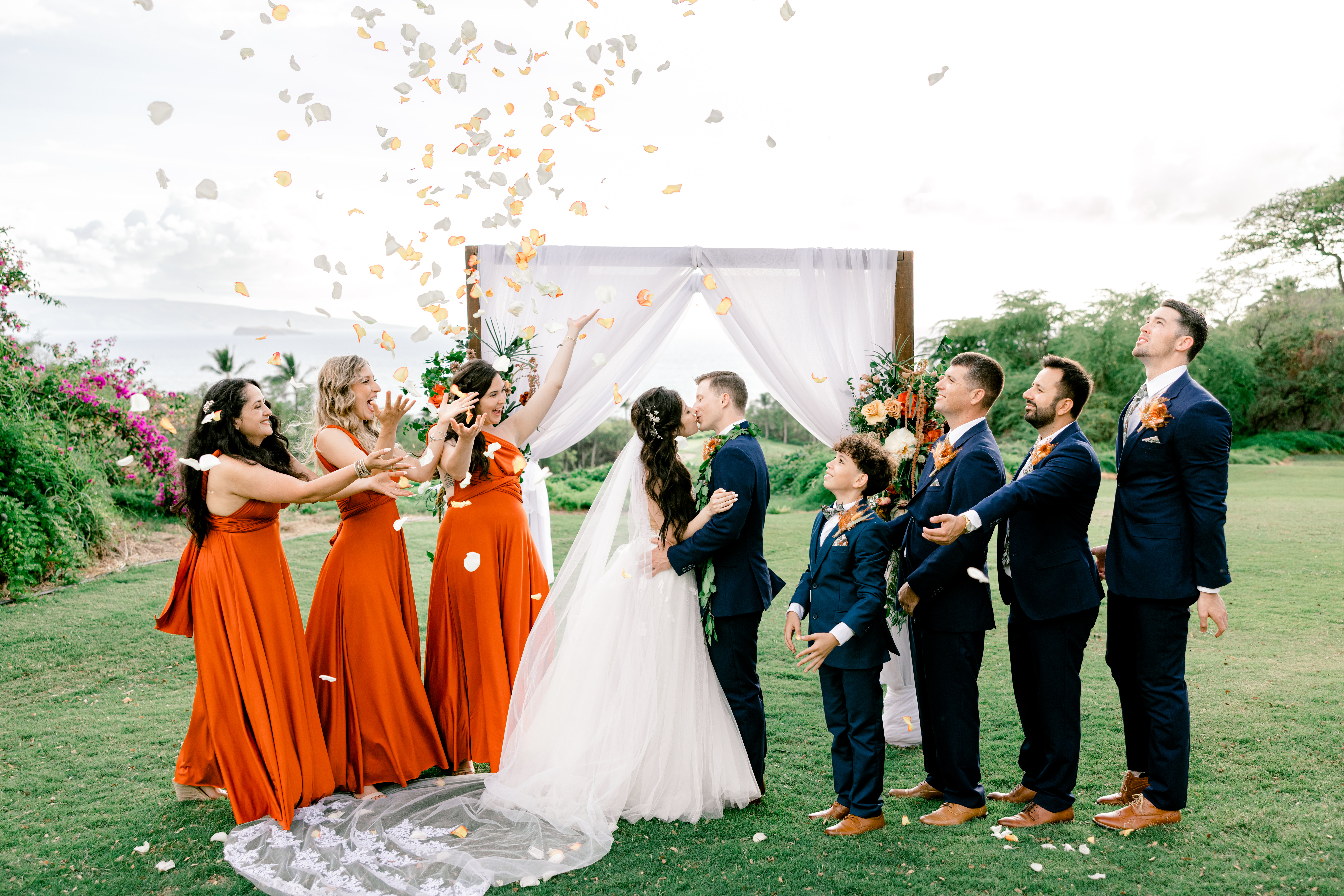 Women wearing wedding dress kissing man wearing suit while bridesmaid wearing orange dresses and men wearing suits throw flowers in the air