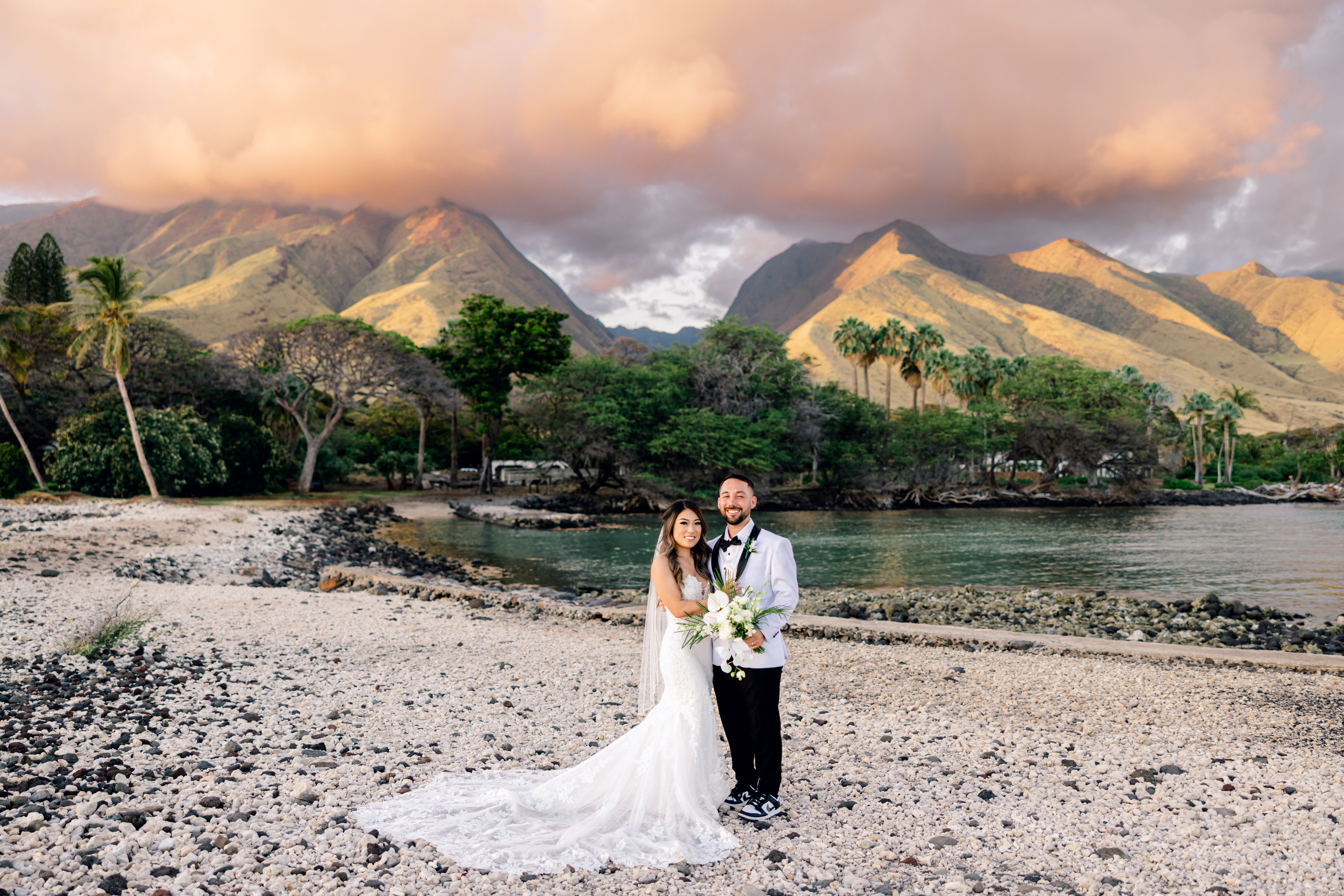Woman wearing white wedding dress standing next to groom wearing black pants and a white blazer on a beach at Olowalu Maui.