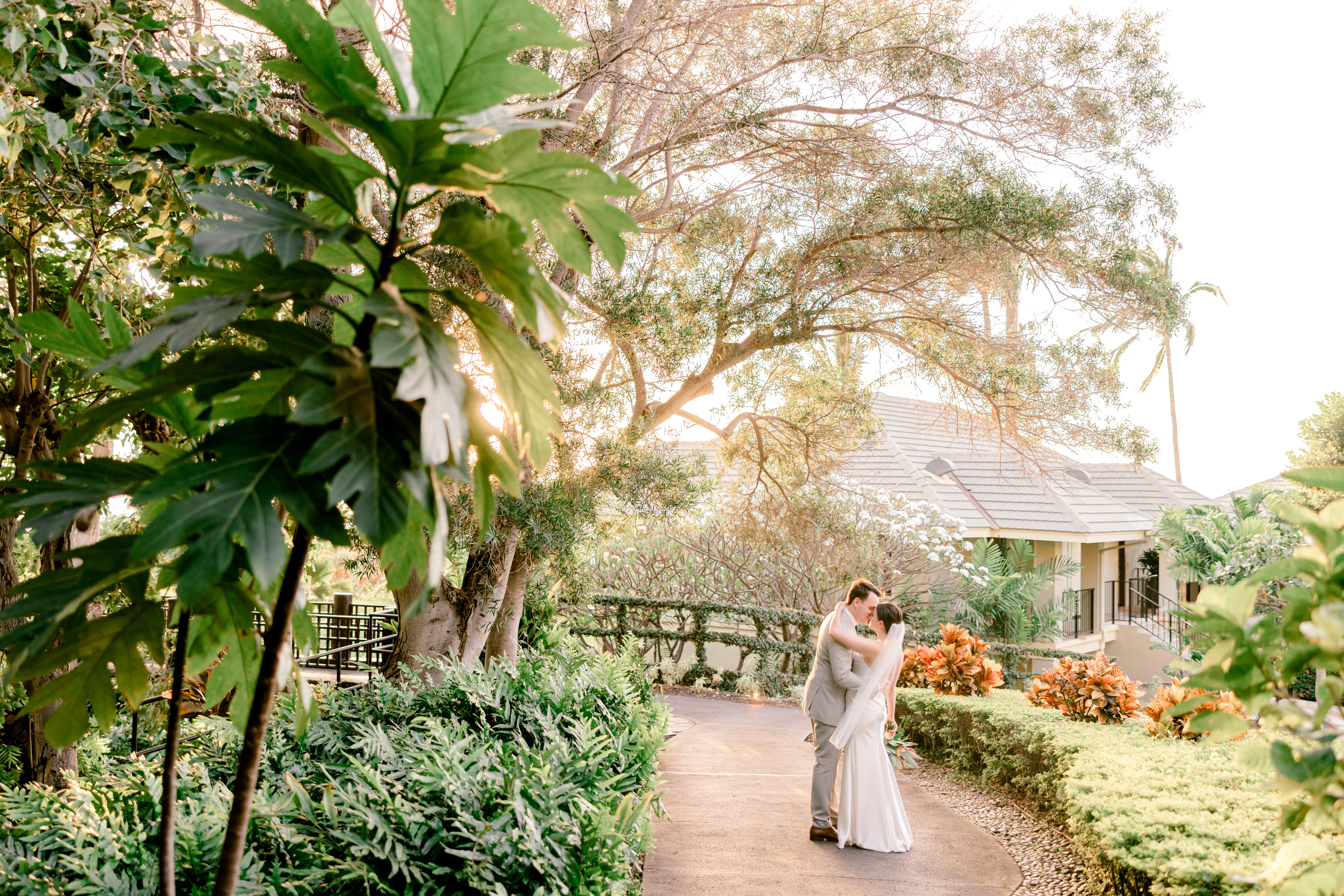 Woman wearing wedding dress hugging man wearing suit while standing under palm trees at Hotel Wailea Wedding Venue