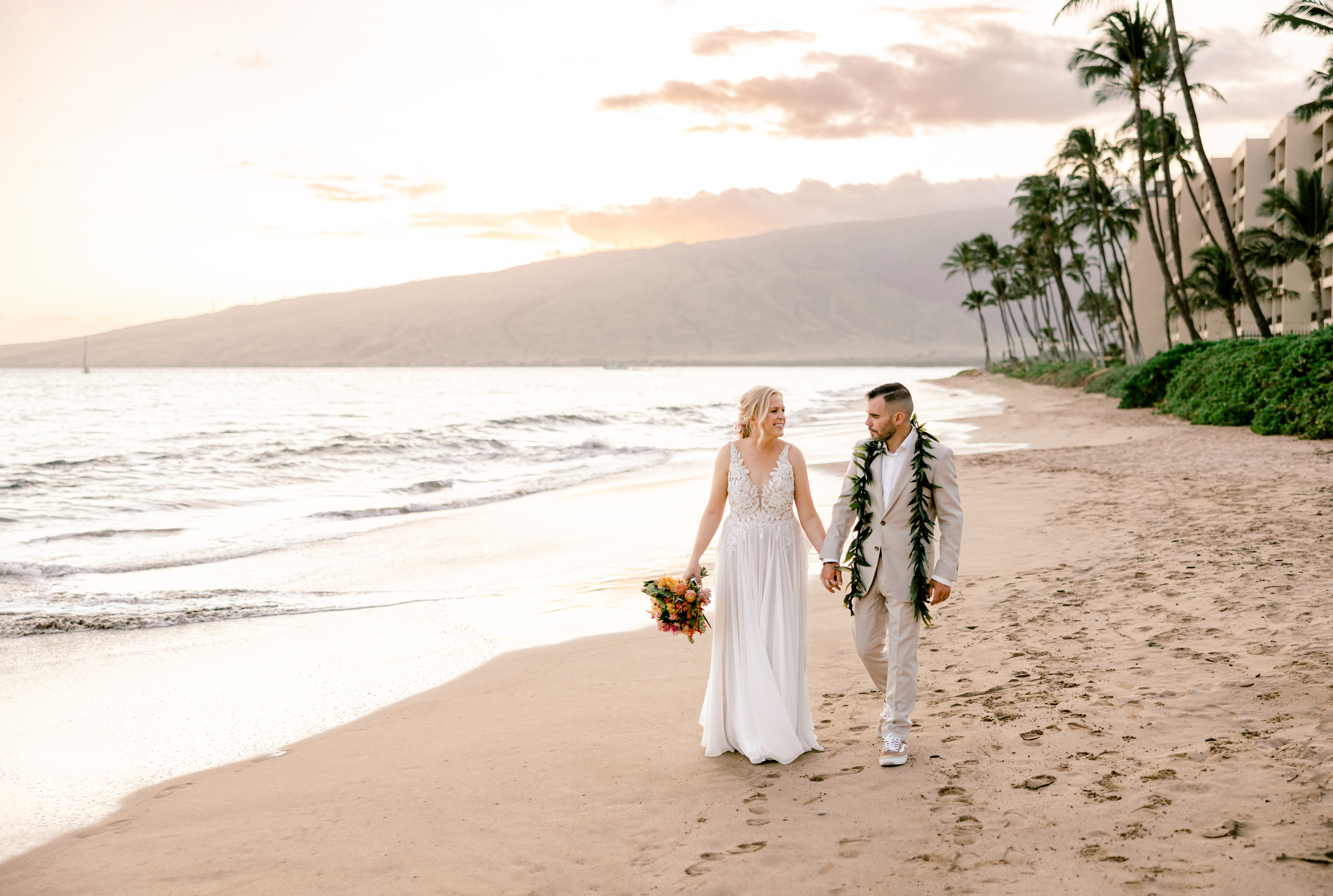 Woman and man holding hands while walking in sand at a beach in Hawaii during sunset.