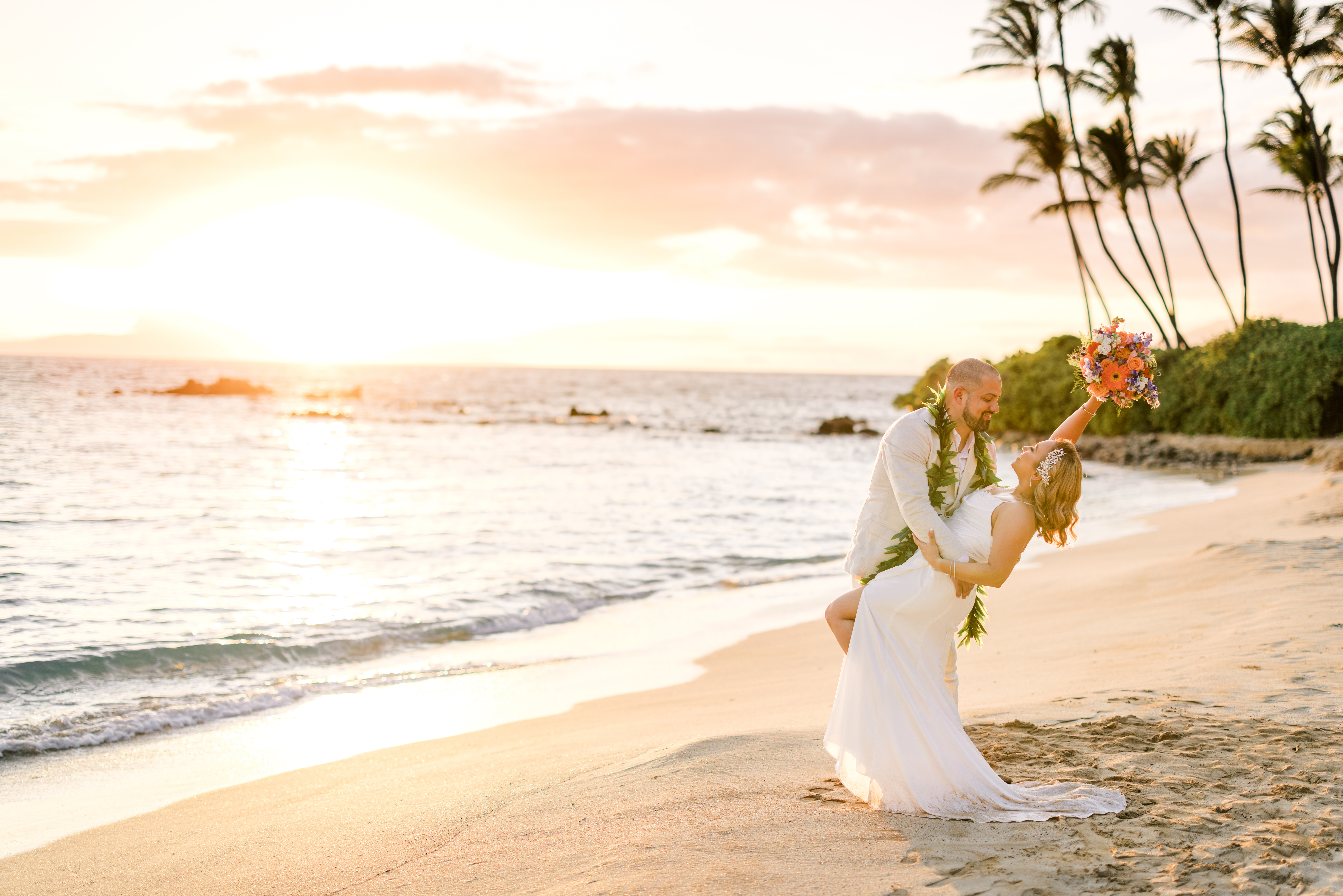 Groom dressing in a suit dips bride wearing white dress and holding flowers in Wailea for their elopement