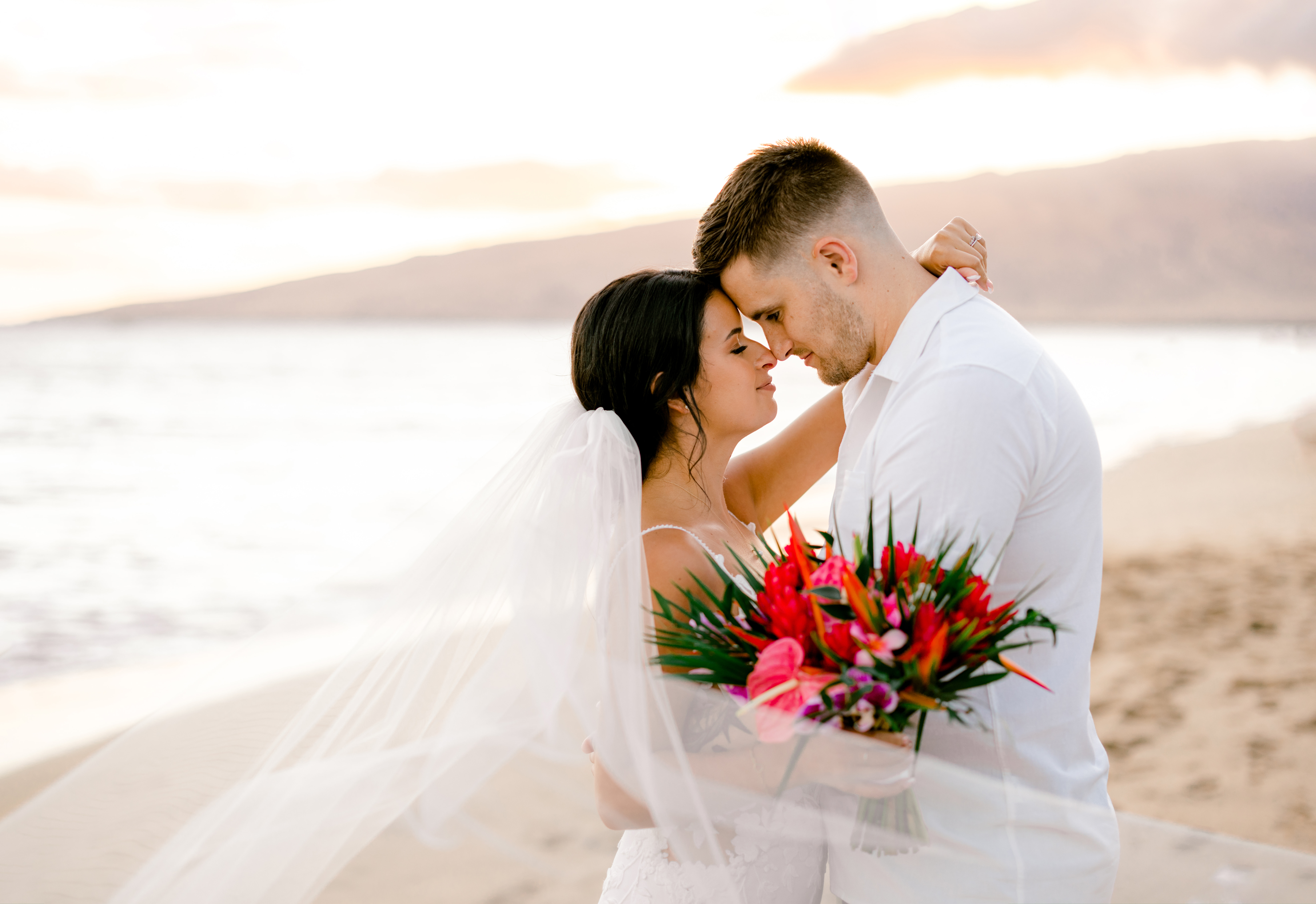 Bride and Groom hugging under bridal veil and holding a hot pink flowers at beach in Kihei, Maui