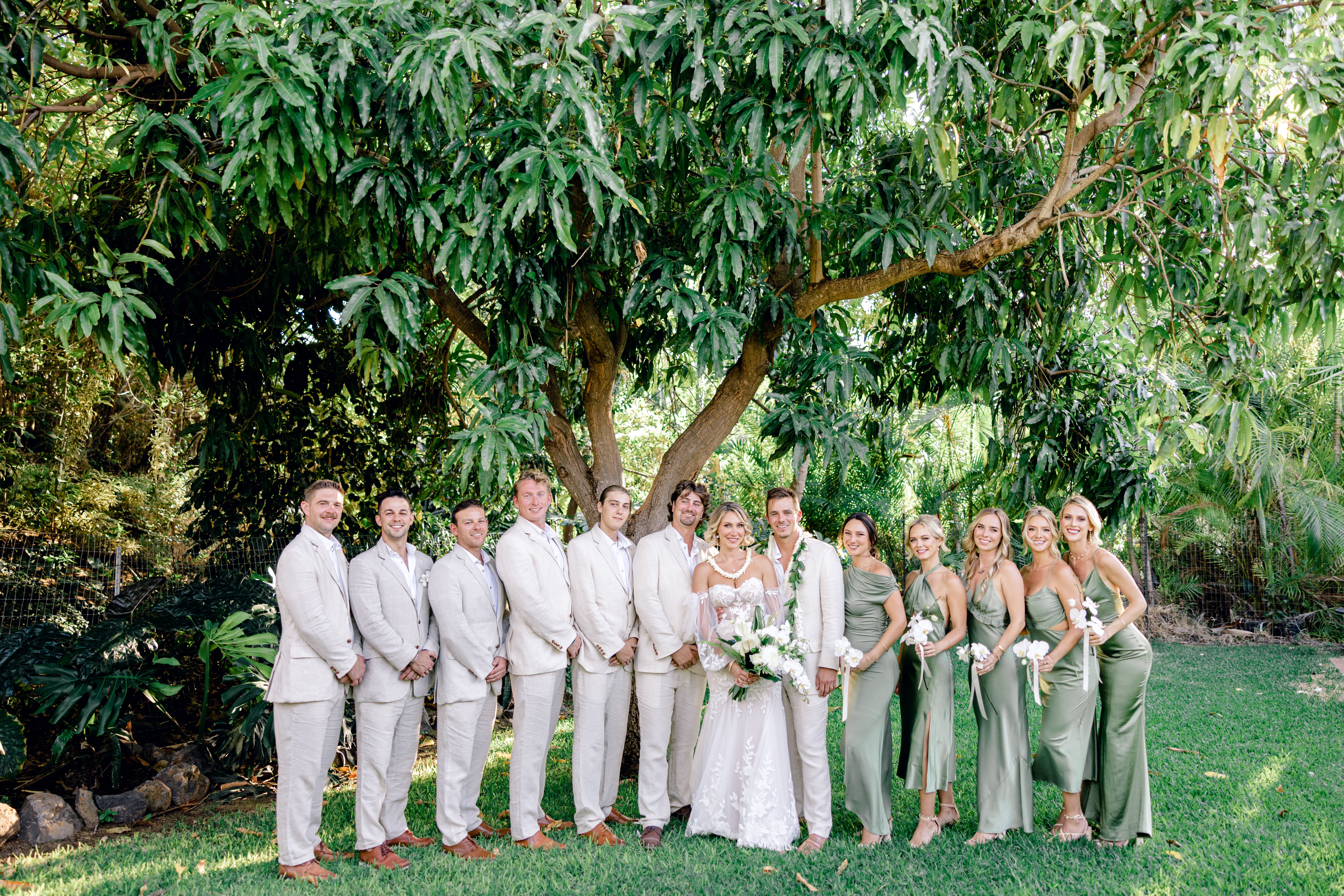 Woman wearing a wedding dress holding flowers and man wearing linen outfit standing beside their wedding party at wedding venue in south Kihei.