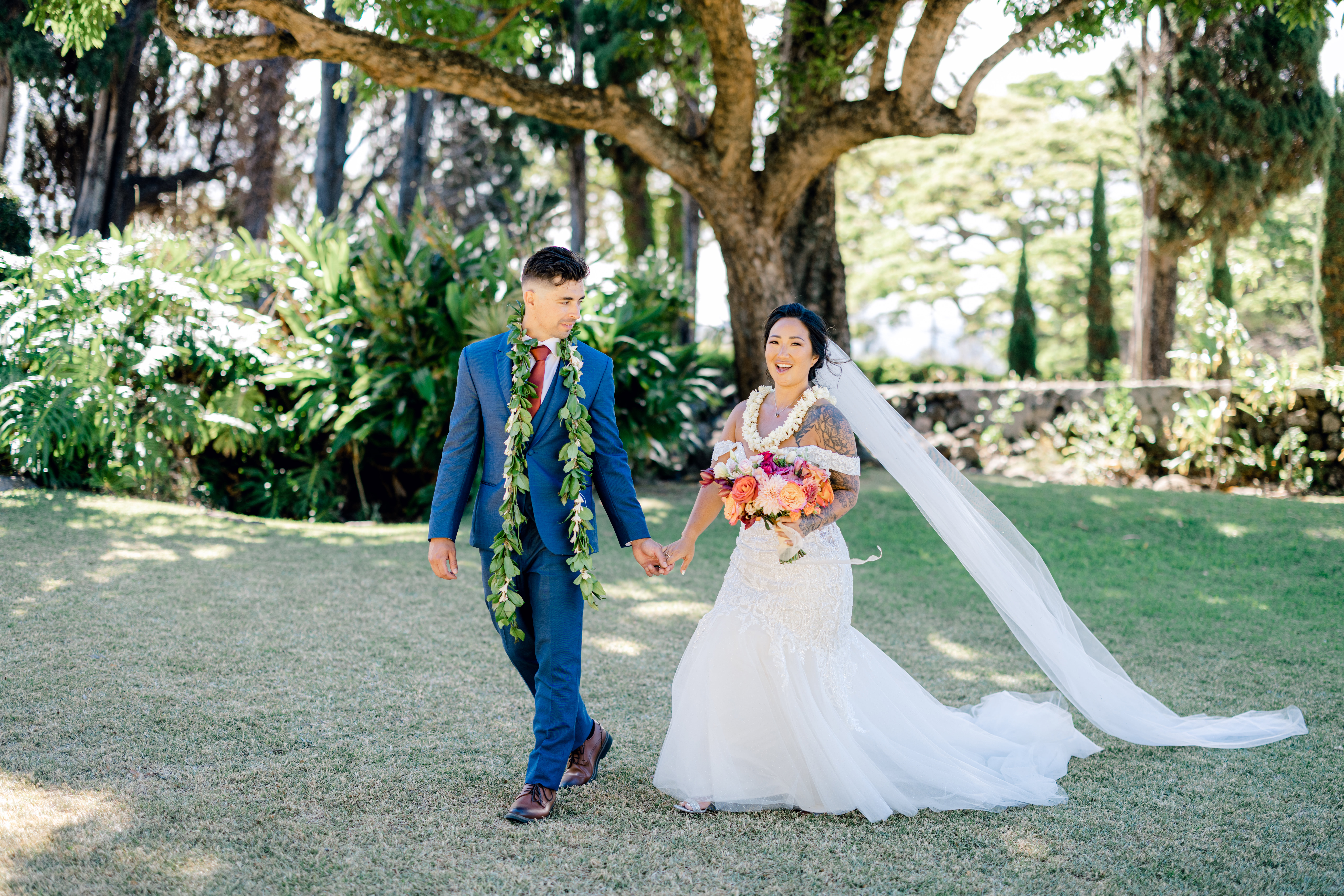 Female wearing a white wedding dress holding a colorful bouquet of flowers while standing next to man wearing a Maile lei looking at woman.
