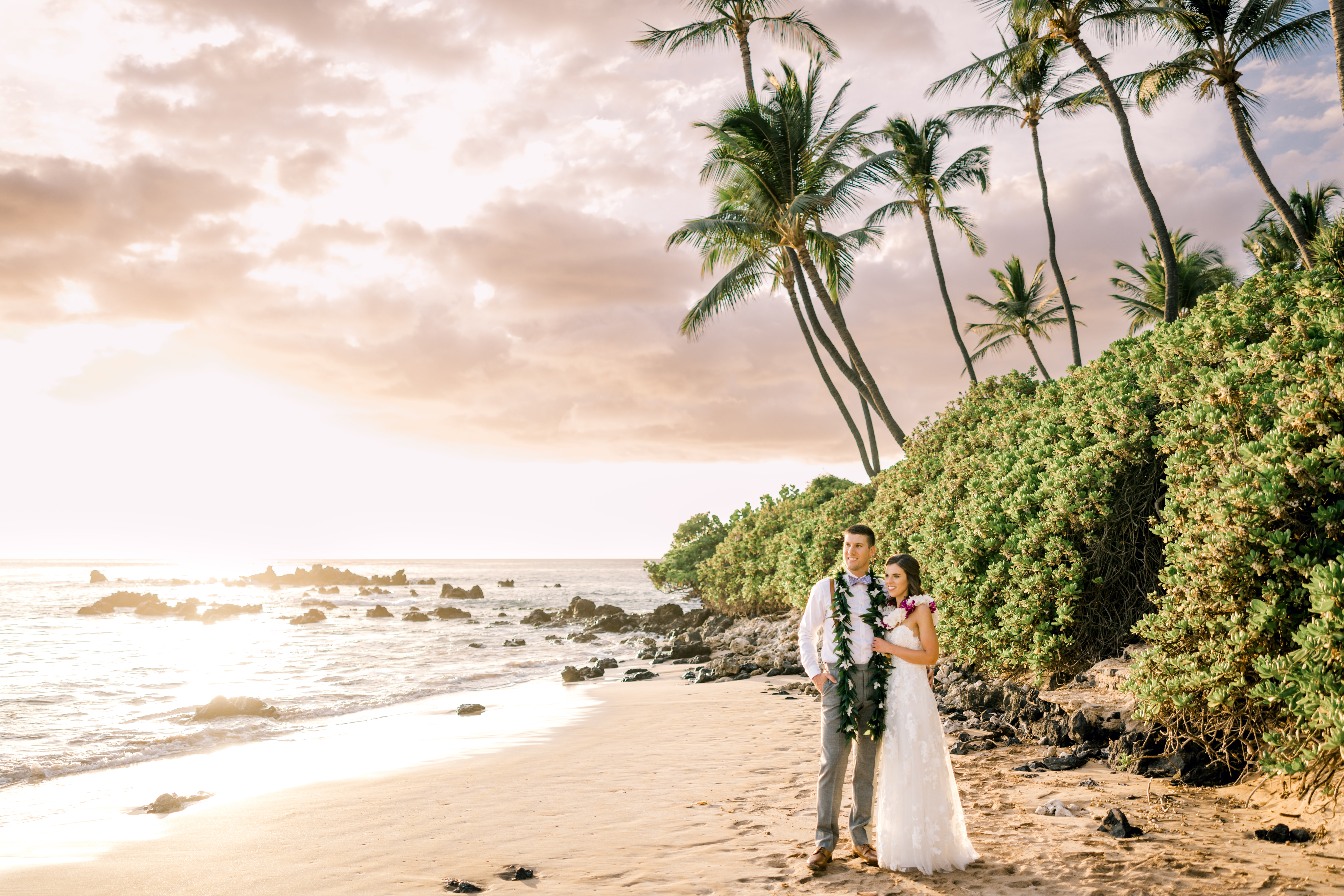 Woman wearing wedding dress and a lei standing next to a man wearing a suit and a Maile lei on a beach in front of Maui's luxury resort