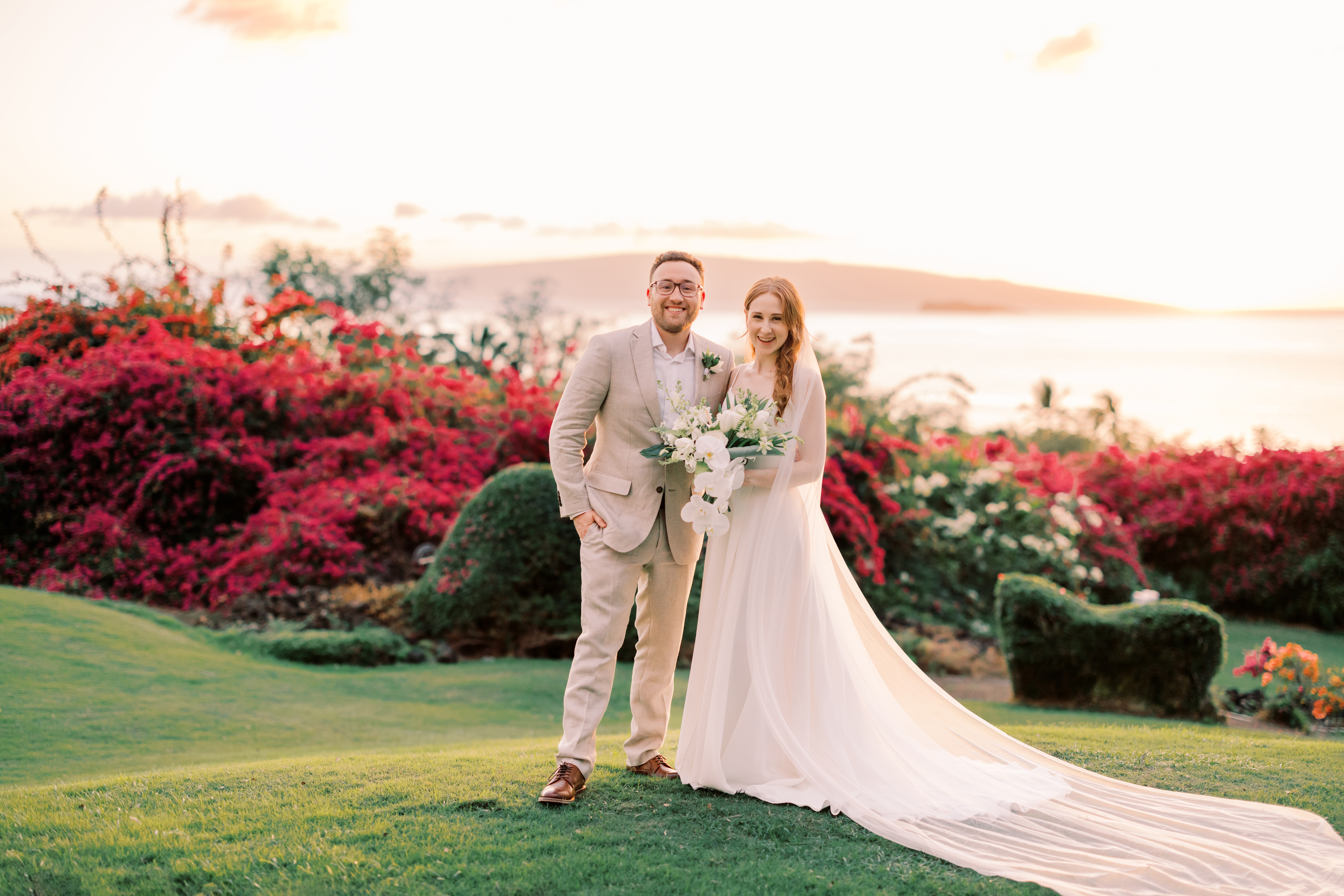 Groom standing beside bride wearing a white wedding dress holding flowers during sunset
