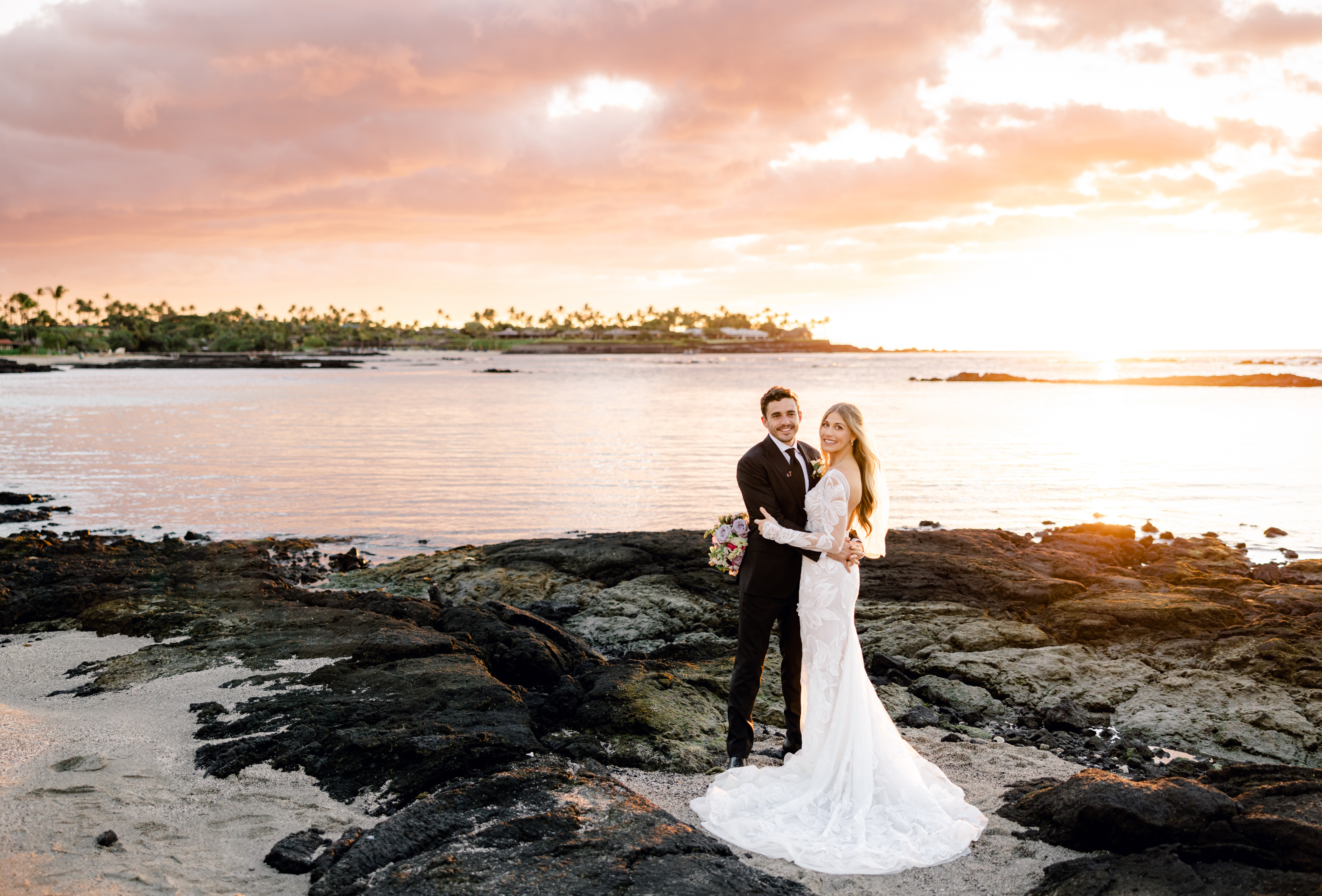 Groom and bride portraits holding slowers are hugging while standing at the beach resort Mauna Lani during sunset.