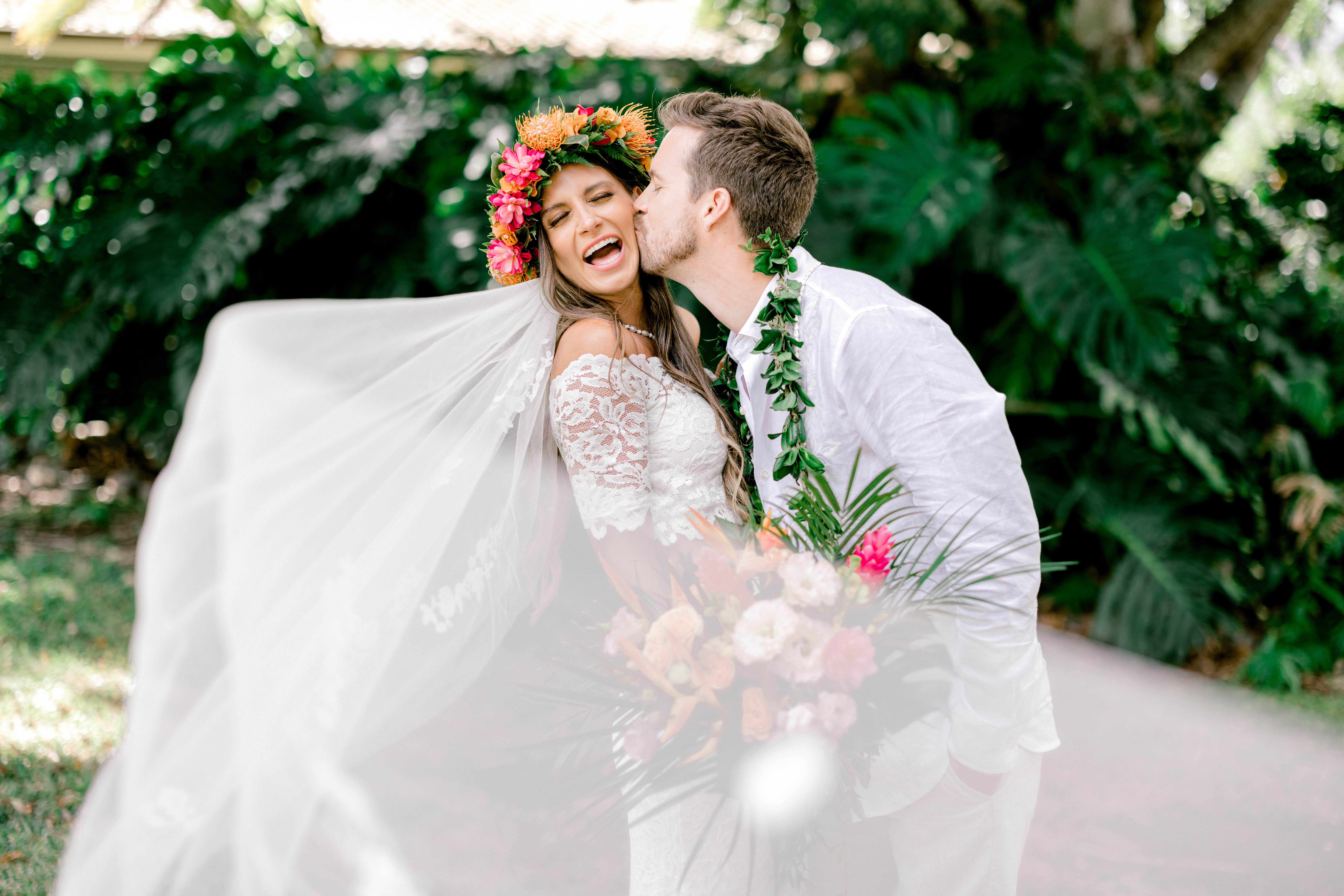 Groom wearing a green maile lei kissing his bride on the cheek wearing a pink flower crown for their wedding portraits in Maui, HI