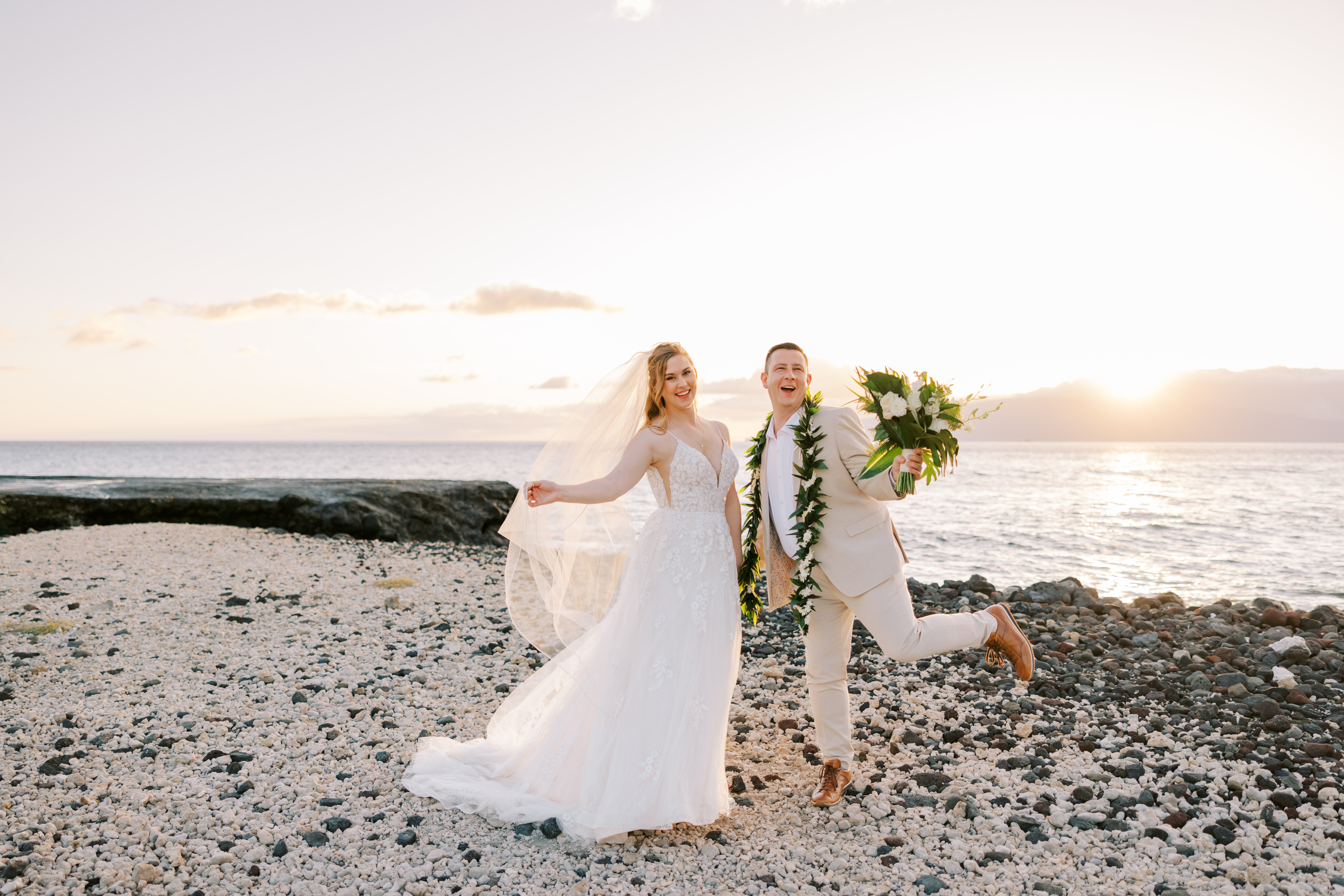 Woman wearing a wedding dress holding hands with a man wearing a brown suit holding flowers during sunset at Olowalu Plantation House.
