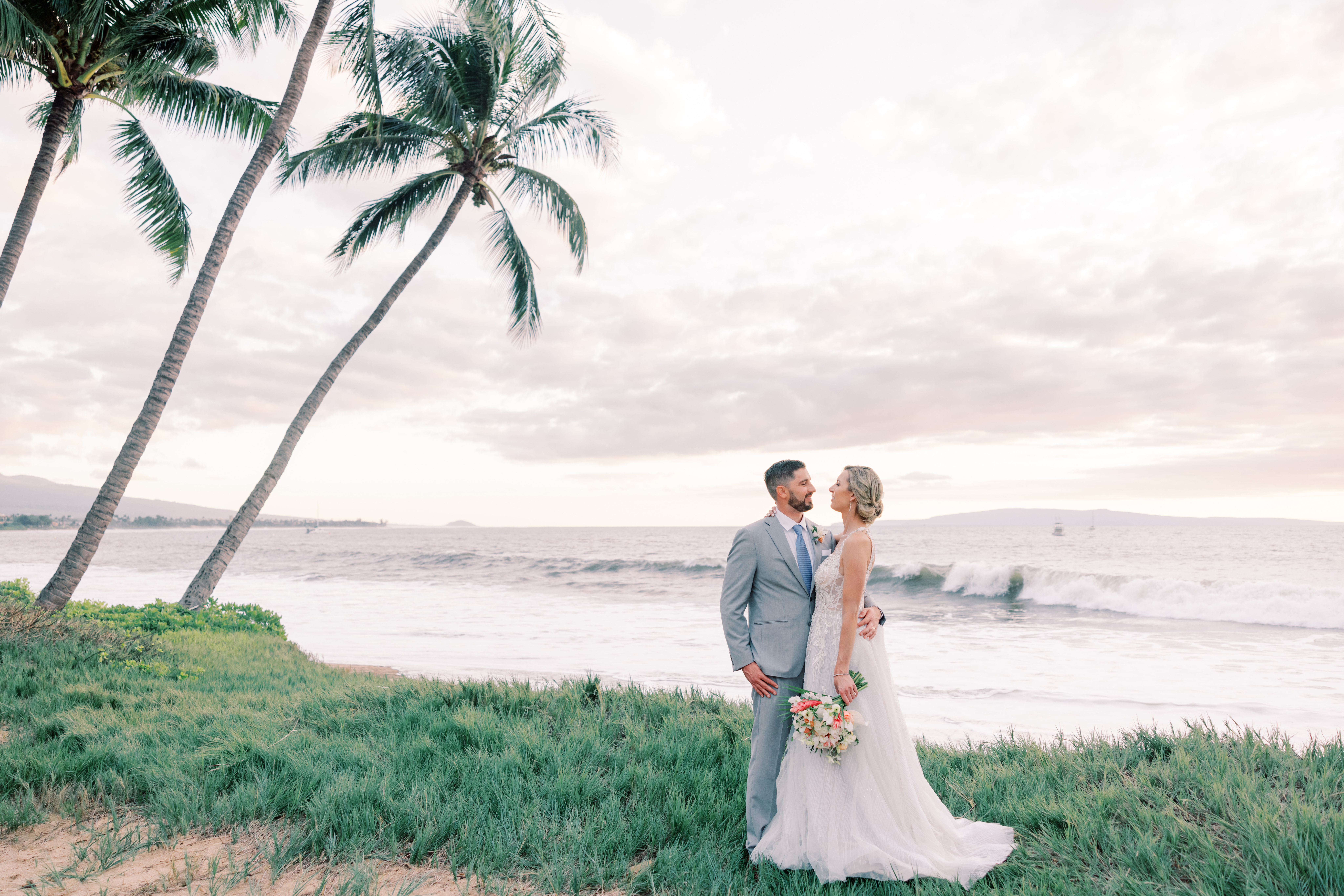 Groom and bride standing at a beach with palm trees and ocean waves in the background.