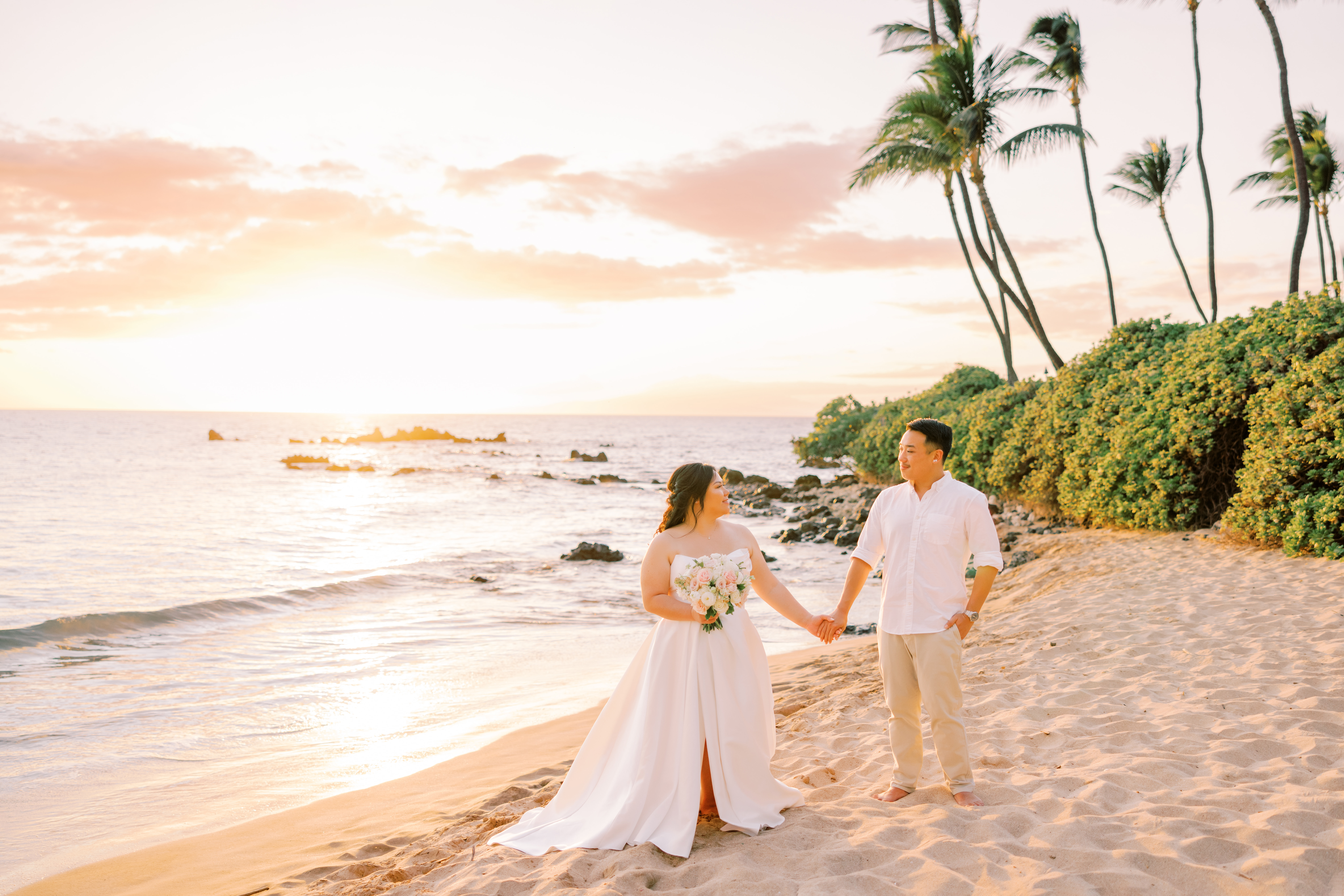 Woman wearing white wedding dress holding flowers and holding hands with a man wearing a shite shirt and brown pants while standing in sand at a beach.