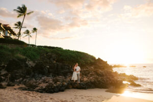 Man and woman hugging standing in front of laval rocks, palm trees, during sunset at a Maui hotel.