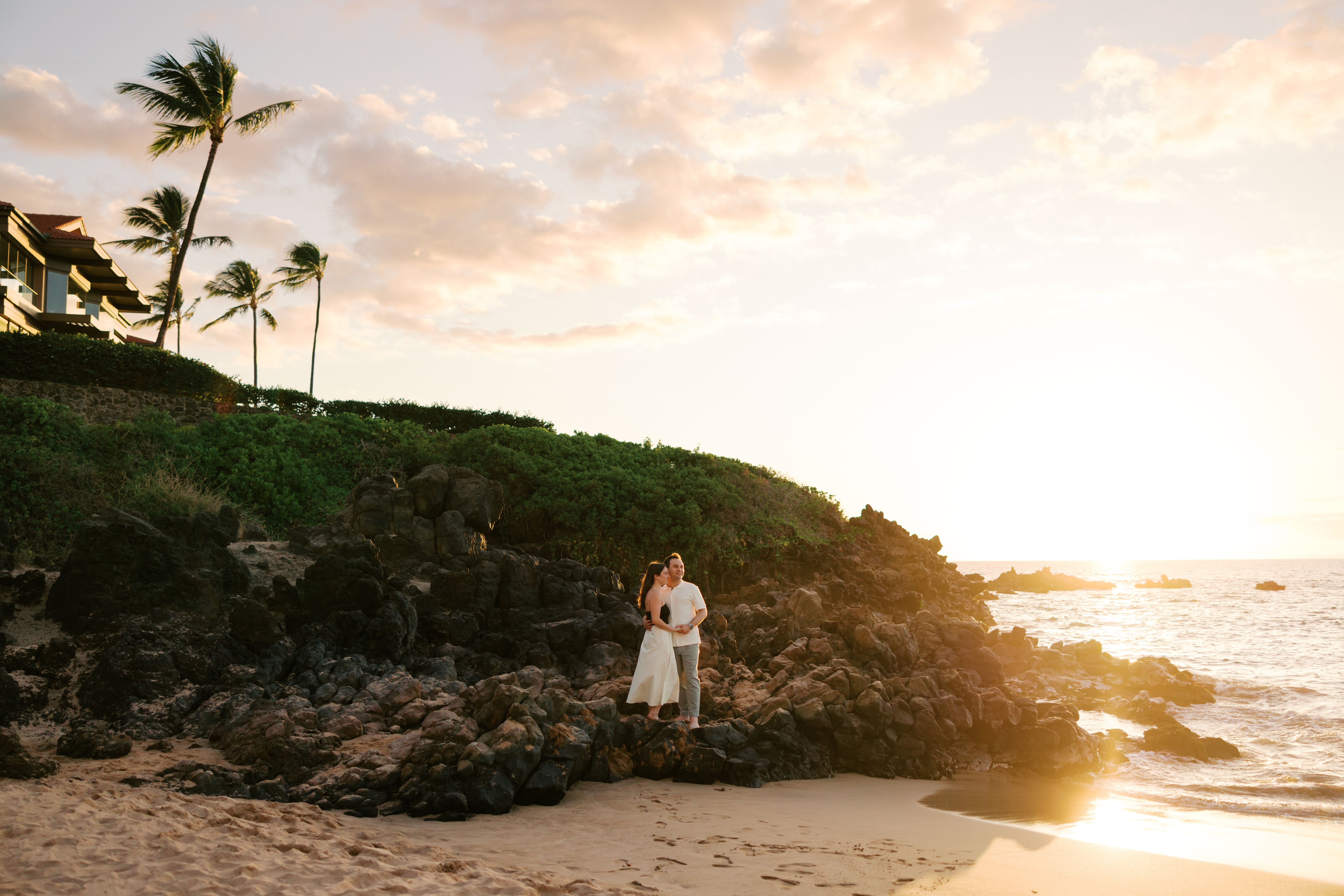Man and woman hugging standing in front of laval rocks, palm trees, during sunset at a Maui hotel.