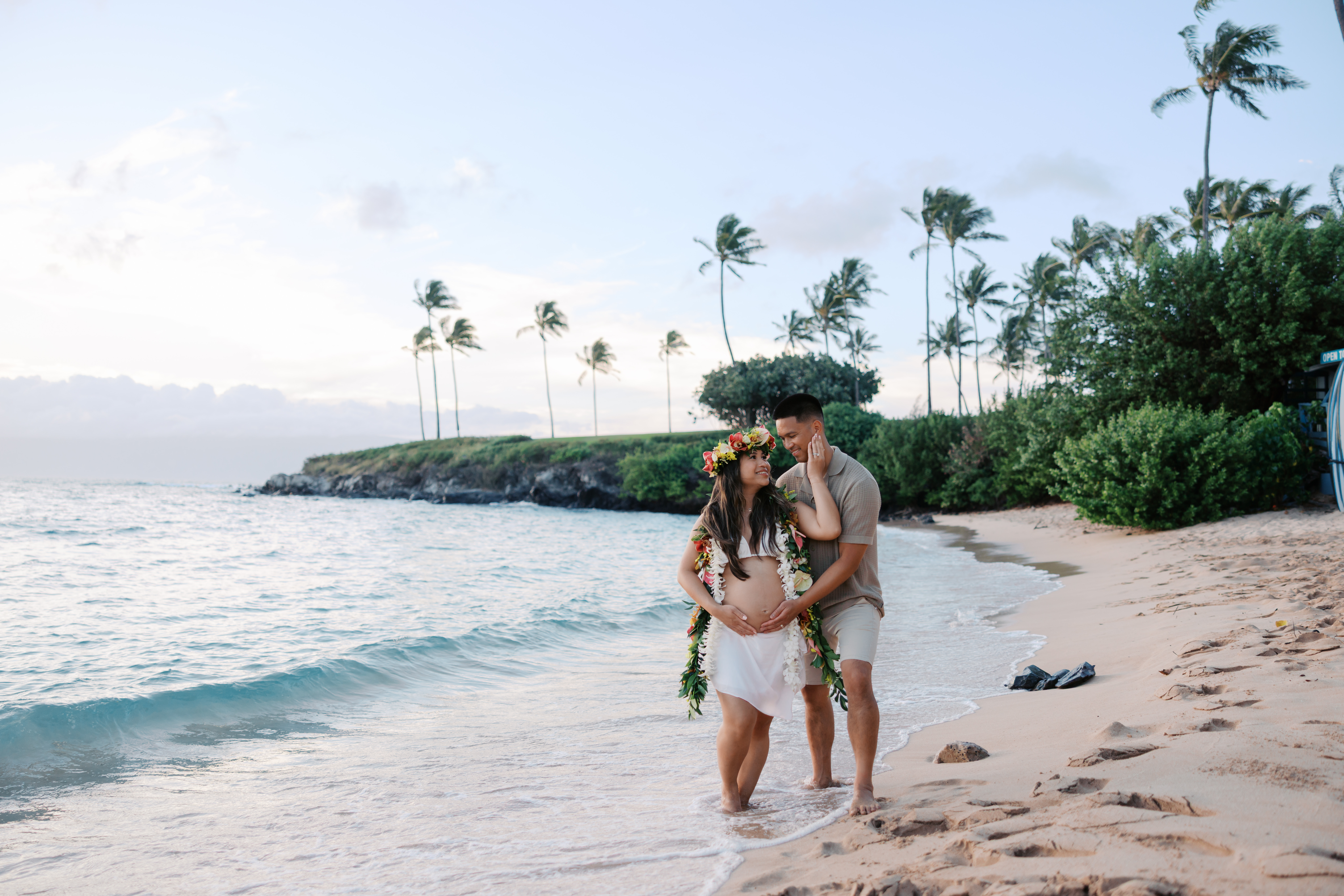 Pregnant lady wearing a flower crown and lei standing next to a man at the beach while man is touching her belly.
