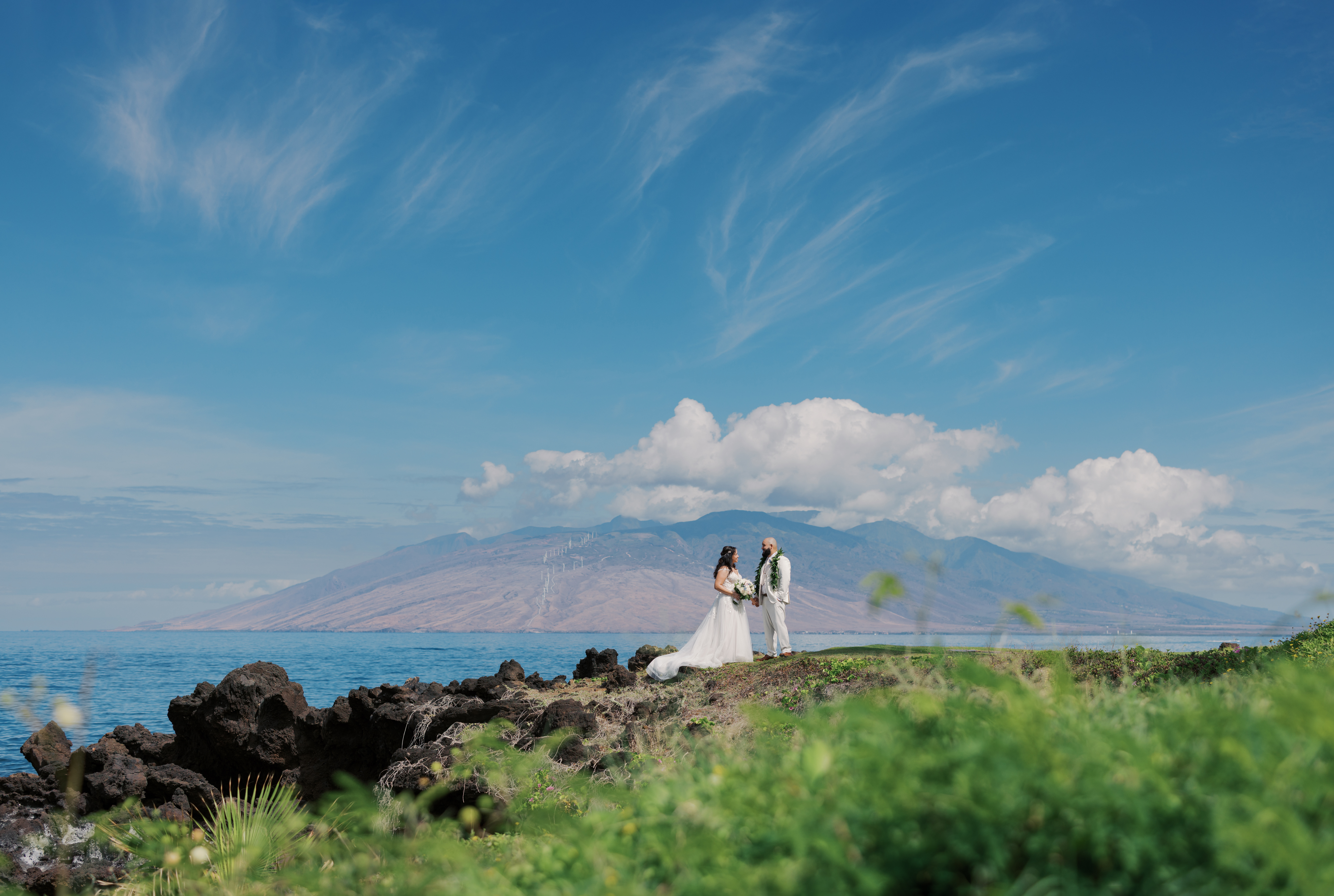 Woman wearing a white wedding dress standing next to a man wearing beige suit on a sunny clear blue sky day at the beach of Wailea Resort in Maui Hawaii