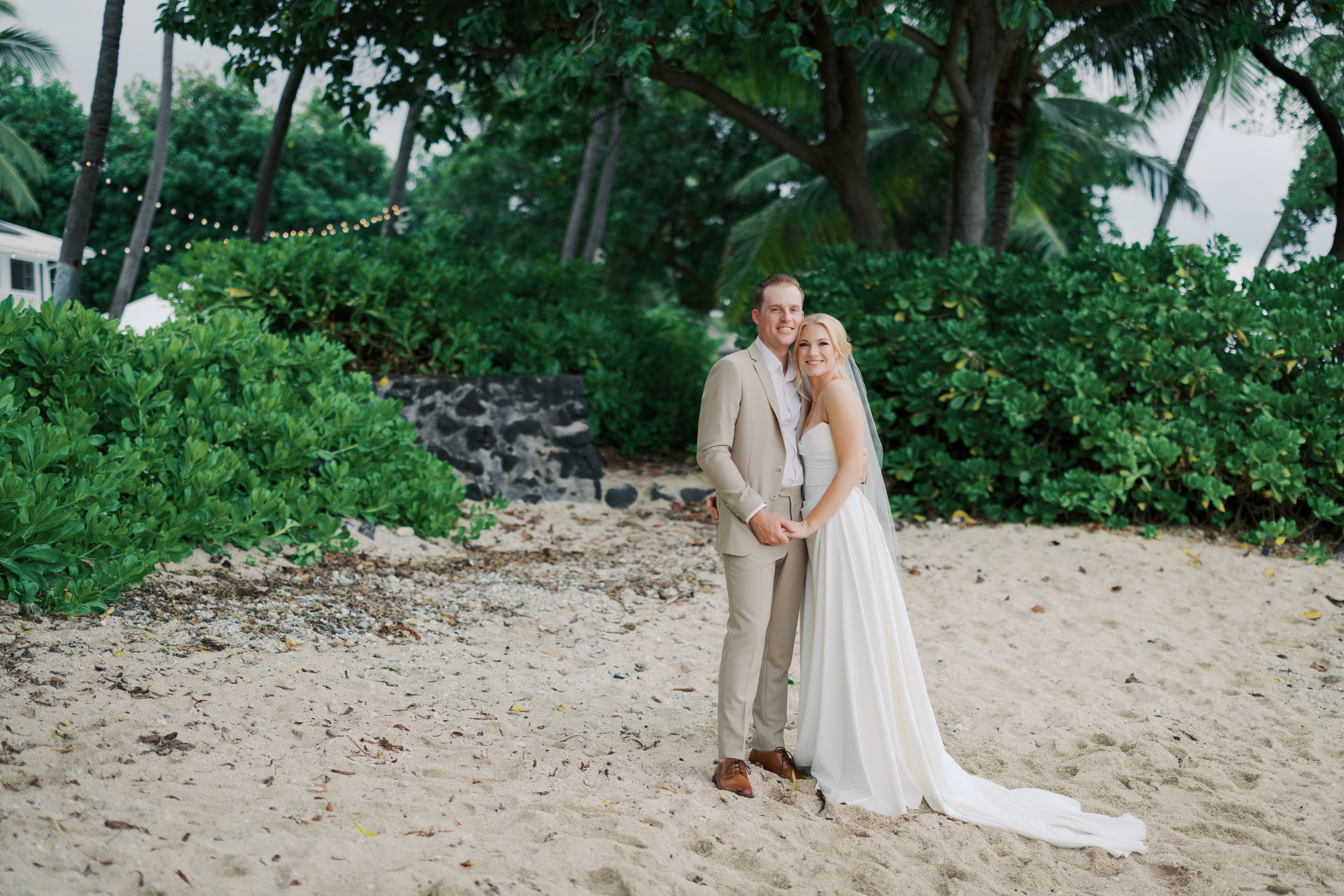 Bride wearing a white dress and viel holding hands with groom wearing a beige suit and brown shoes on a beach in Kona Hawaii with green bushes in the background