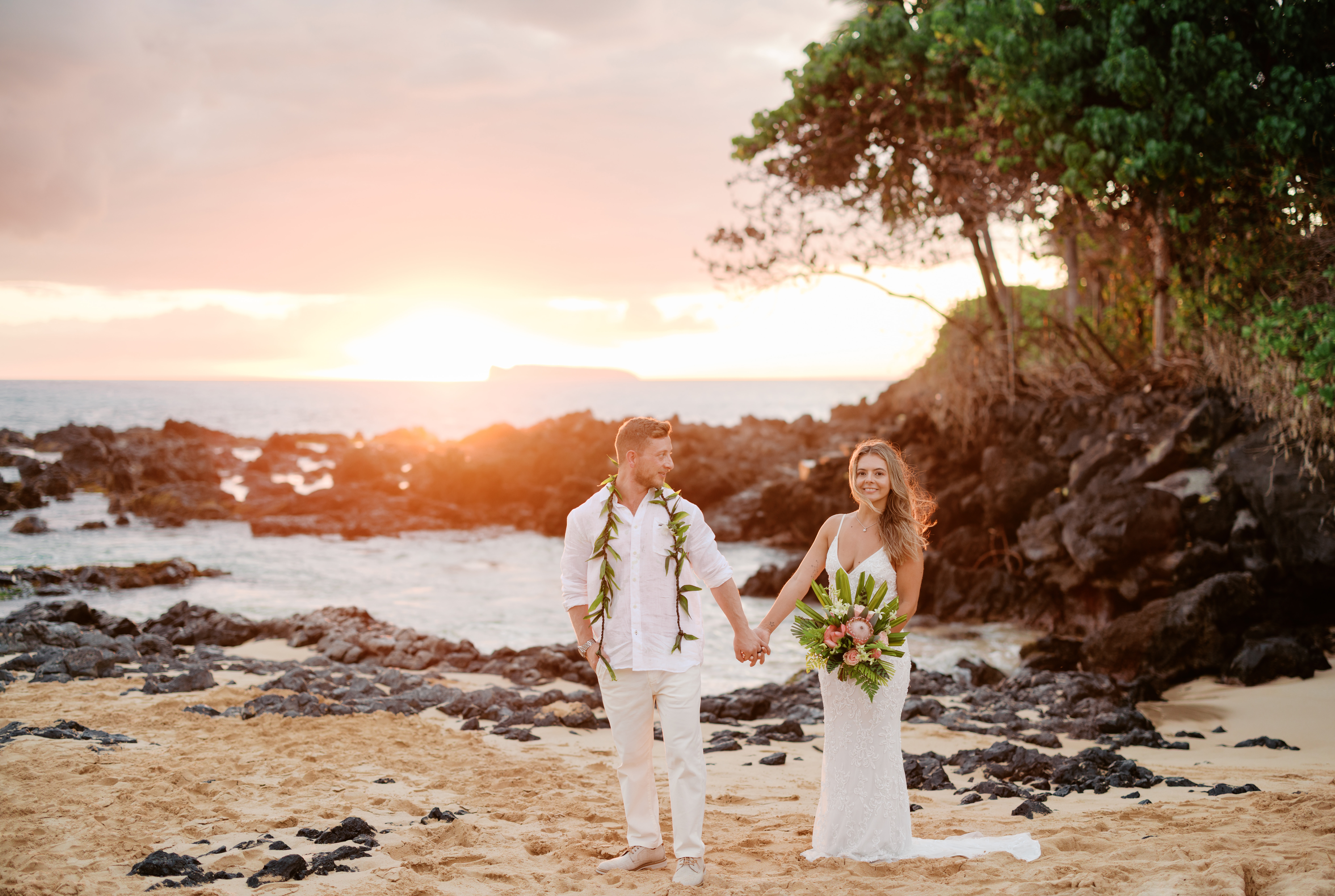 Groom who has dark brown hair wearing a green ti leaf lei holding hands while looking at bride who has long blond hair wearing a white wedding dress holding bouquet of flowers standing in the sand on a beach during sunset