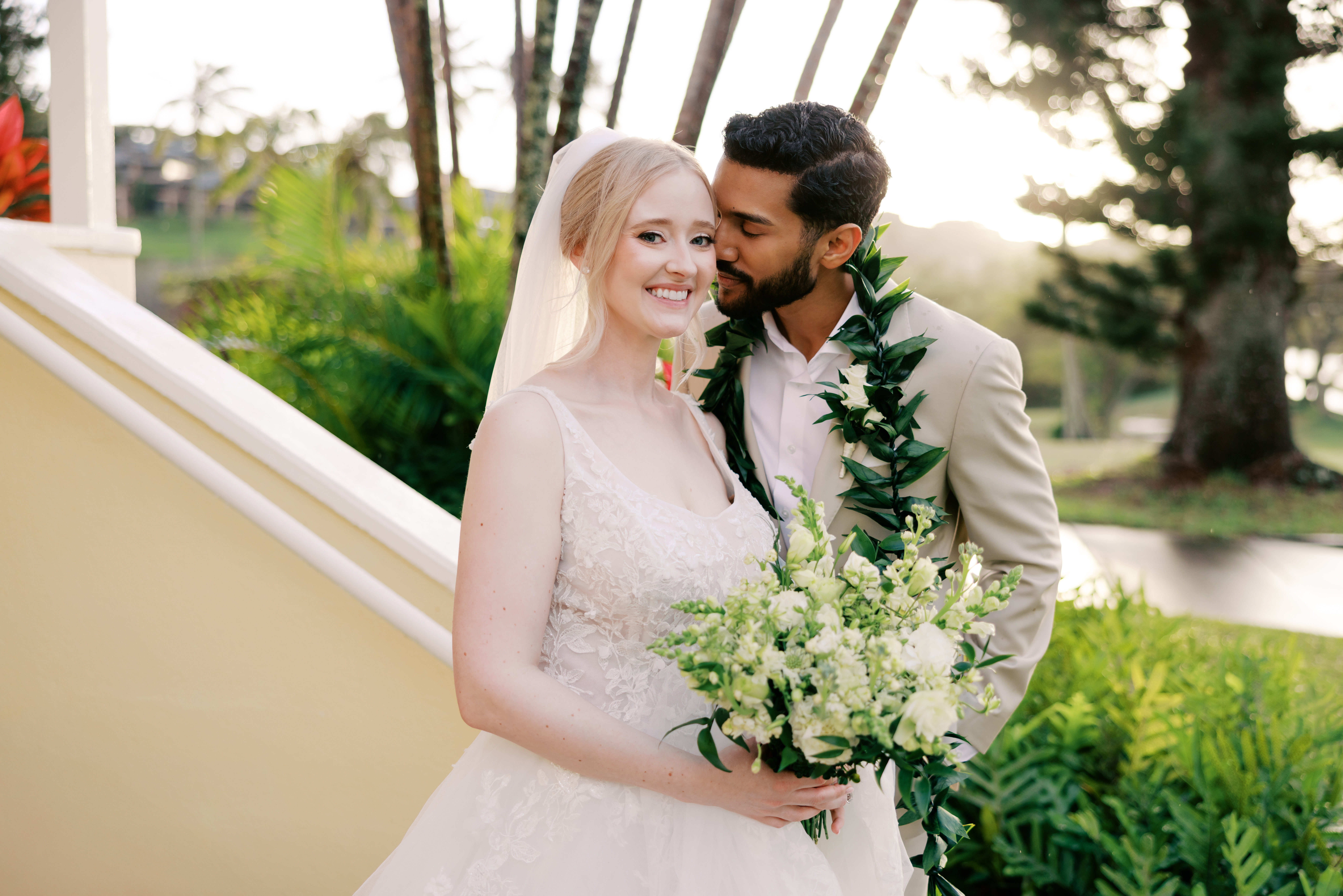 Blong haired Woman wearing a white wedding gown being kissed by her dark haired husband man who is wearing a maile lei.