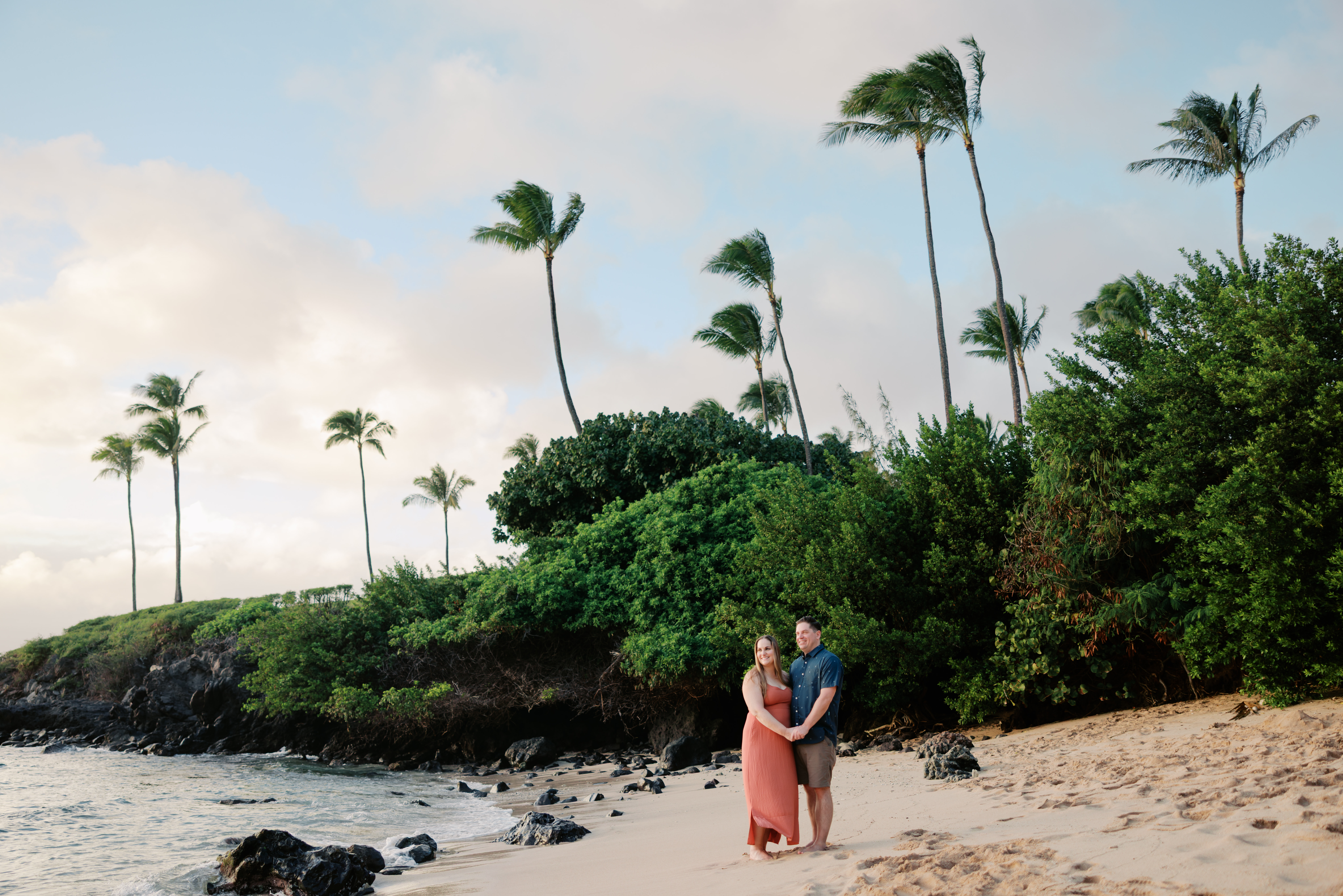 Man and woman holding hands while standing in the sand at the beach in front of the green palm trees.