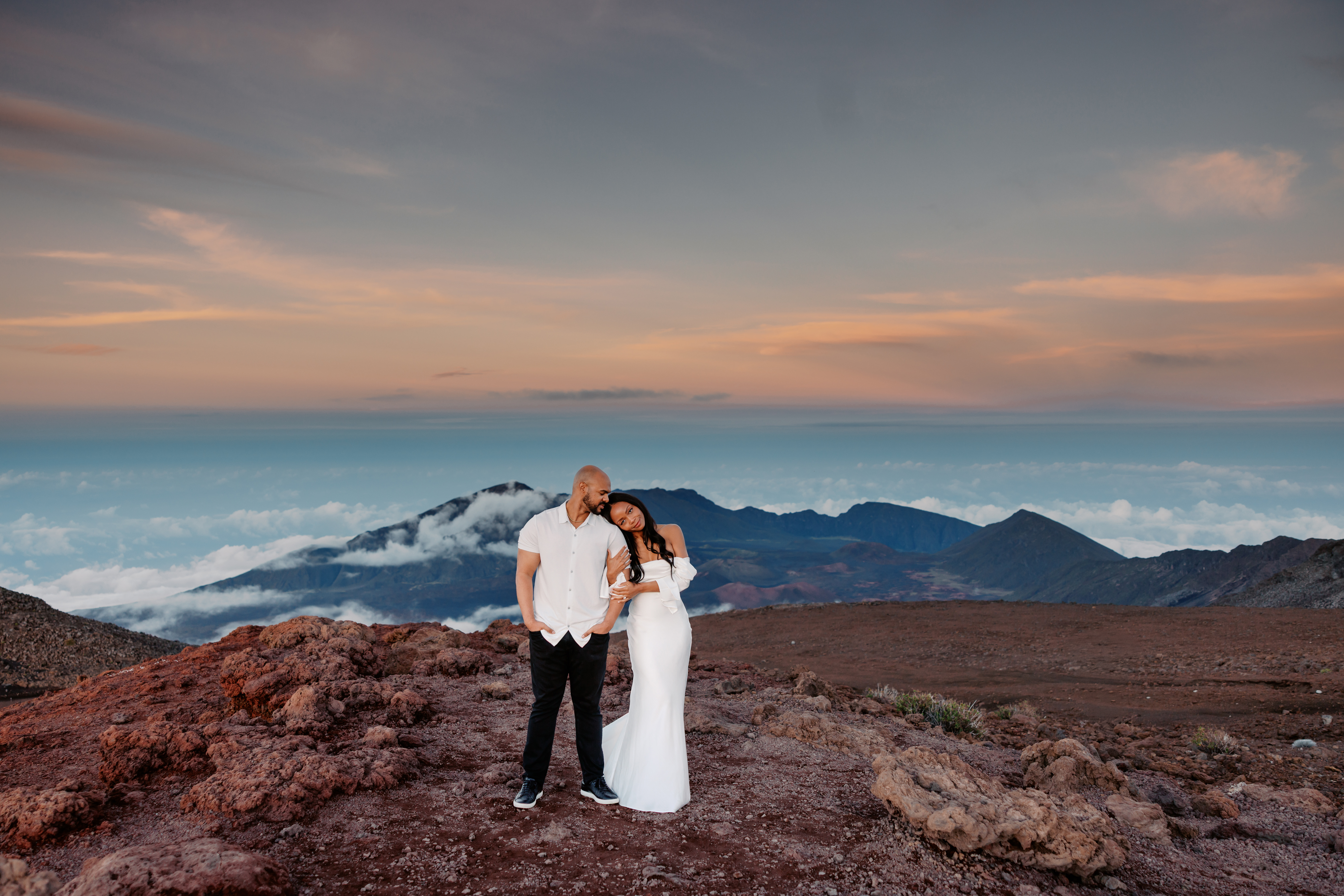 Woman with long black hair wearing a long fitted white dress laying her head on mans shoulder in the evening at Haleakala Park, Maui Hawaii