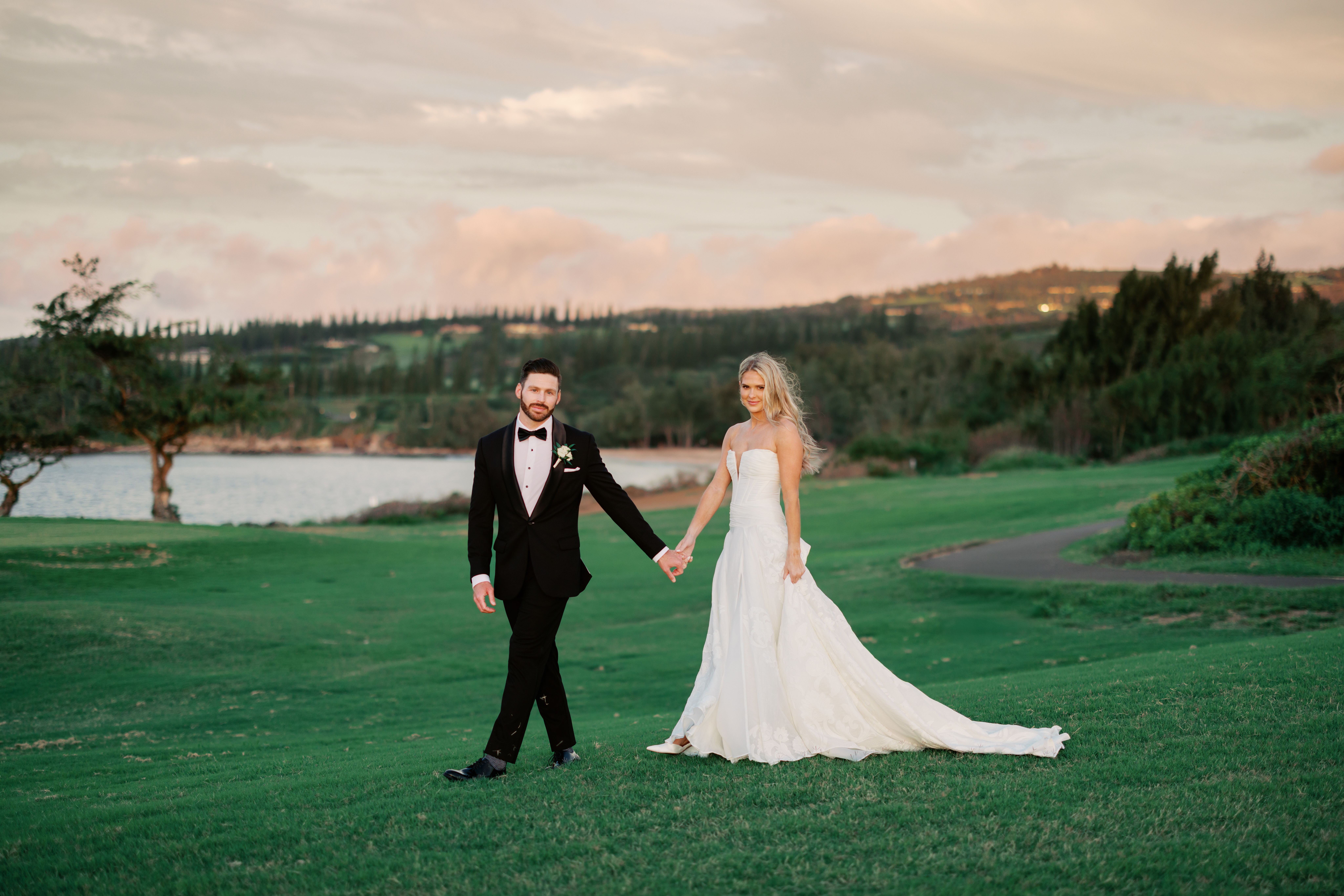 Woman with blond hair wearing a white wedding dress holding hands with a black haired man wearing a black tux standing on the gold curse at Ritz Carlton resort in Maui, Hawaii