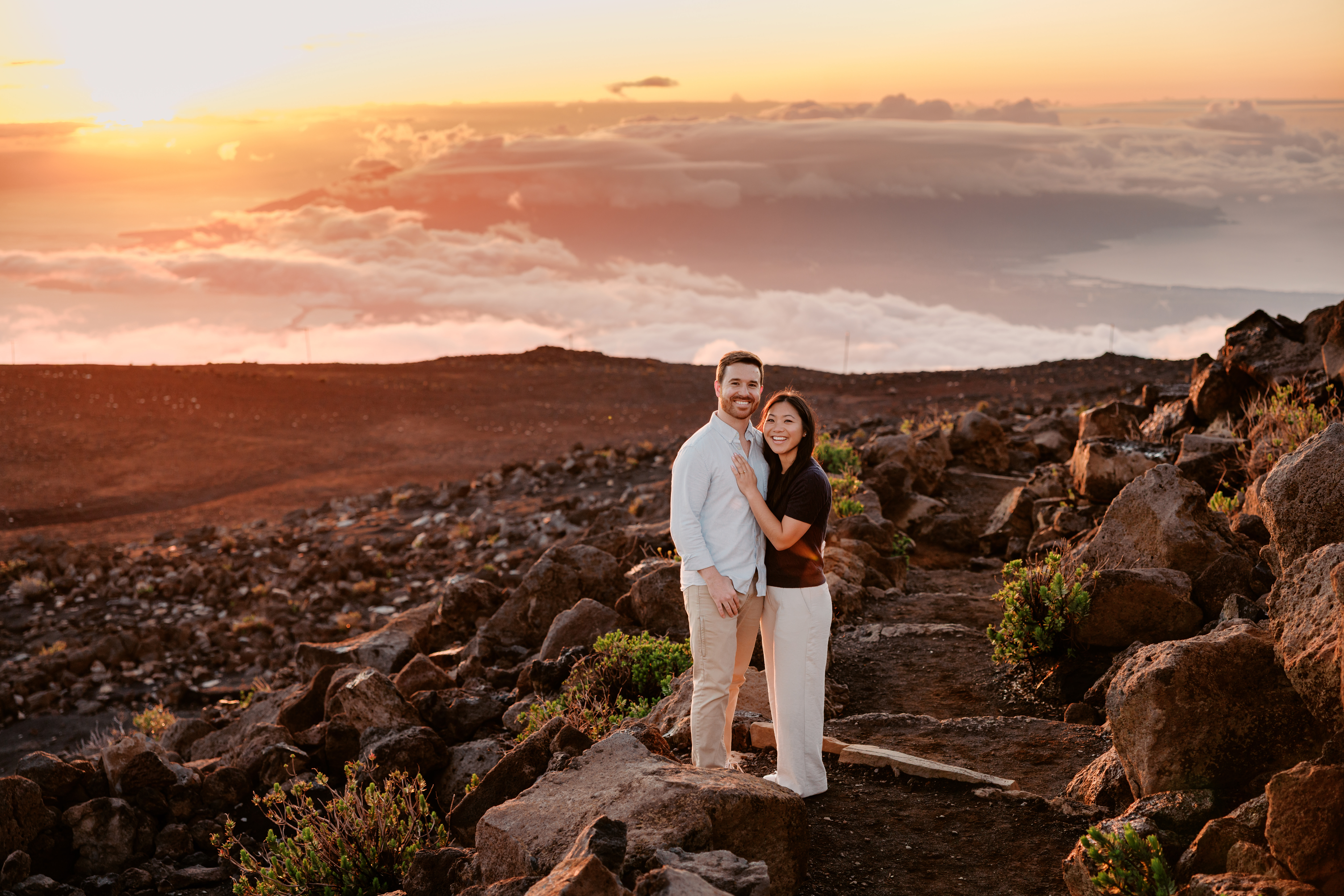 Black haired woman and Brunette man standing at Haleakala Park with sunset, clouds, and red rocks in the background.