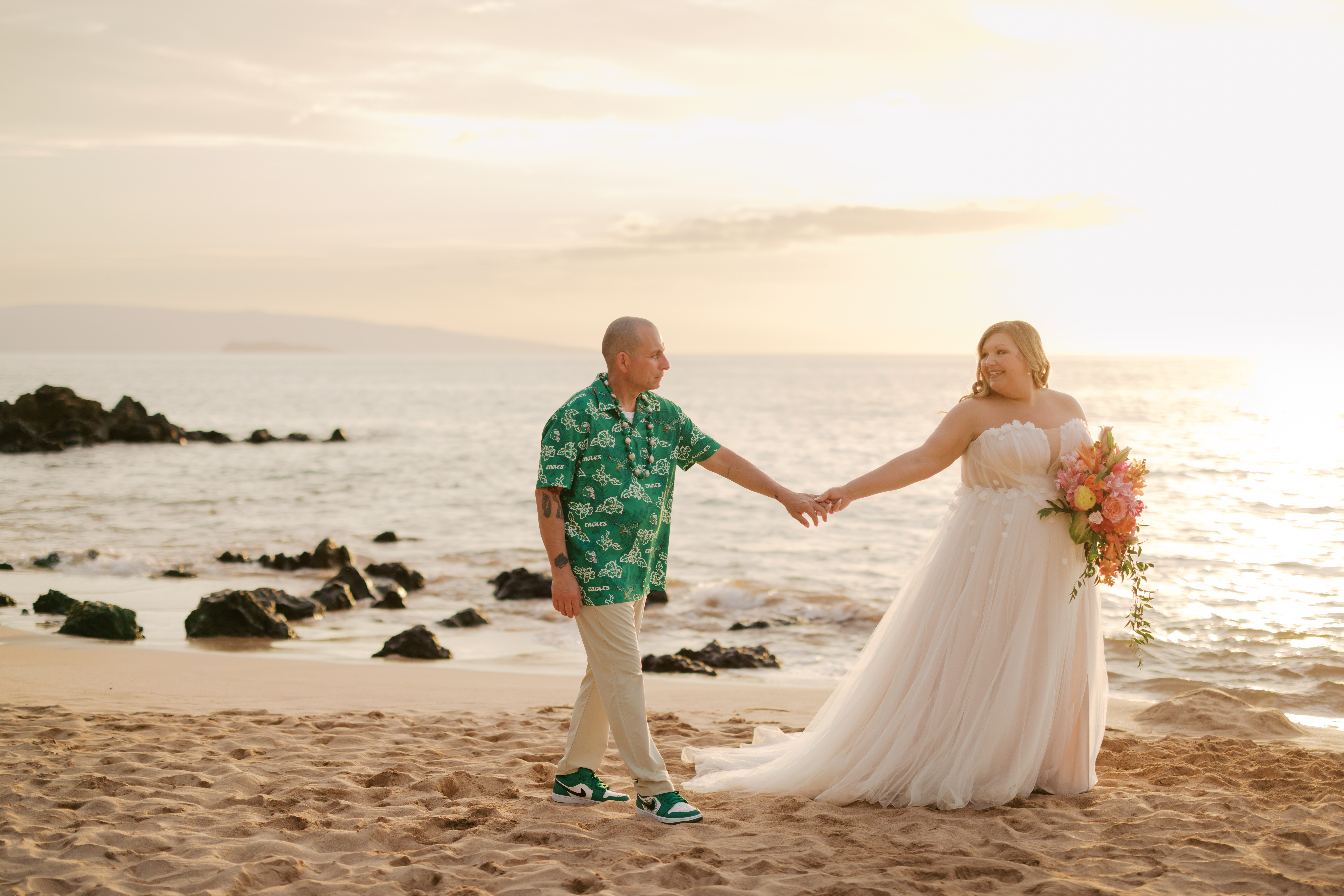 Woman wearing a wedding dress and holding tropical pink flowers holding hands with a man wearing a green tropical shirt during sunset on a beach in Maui, HI