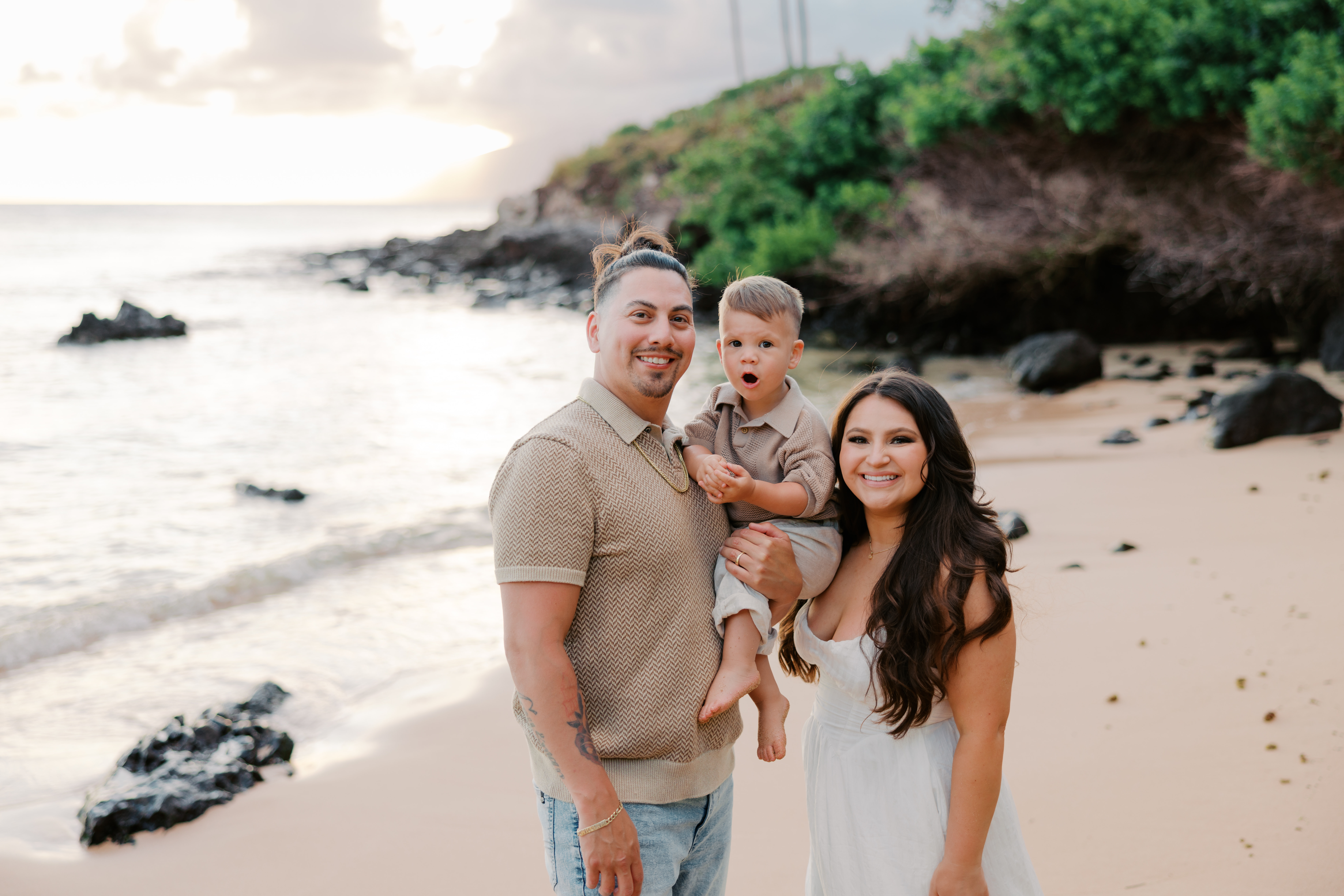 Man smiling holding his baby boy standing next to woman with long dark hair smiling at the beach in Kapalua, Maui, Hawaii.